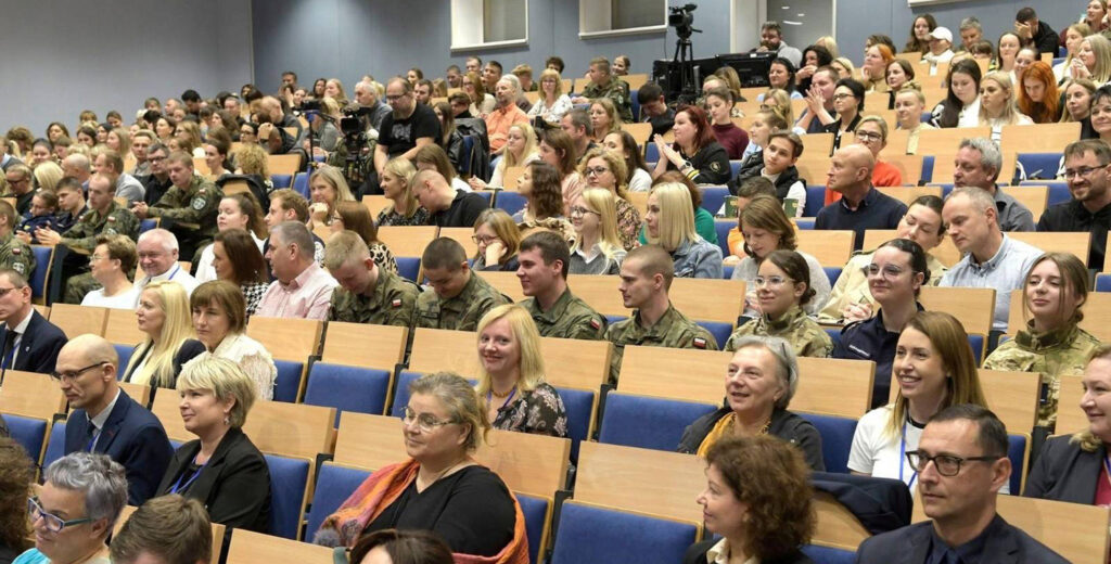 Ludzie siedzą w auli i słuchają wykładu. W tle sprzęt wideo do transmisji spotkania w internecie.